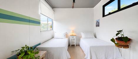 A small, clean room with two beds, a nightstand, a lamp, a potted plant, and a window.