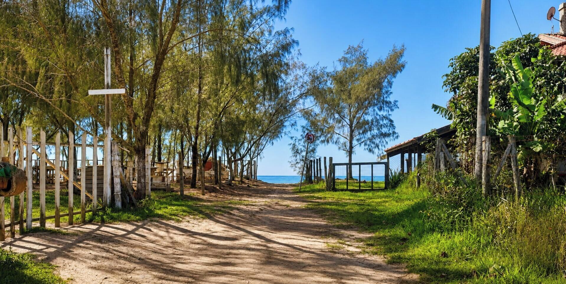 Un sentier de terre mène à une plage, avec une clôture d’un côté et une structure en bois de l’autre.