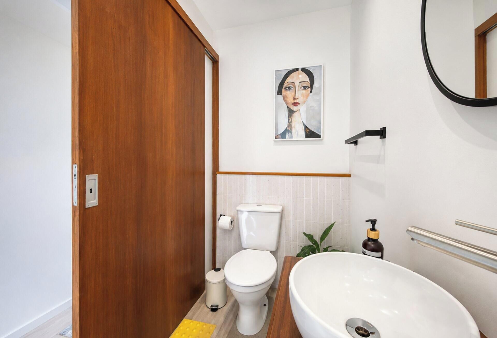 A bathroom with a wooden door, a toilet, a sink, a mirror, and a painting on the wall.