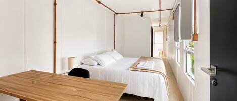Une pièce minimaliste comprenant un lit, une table en bois et une chaise. Une fenêtre offre une vue sur la verdure environnante.