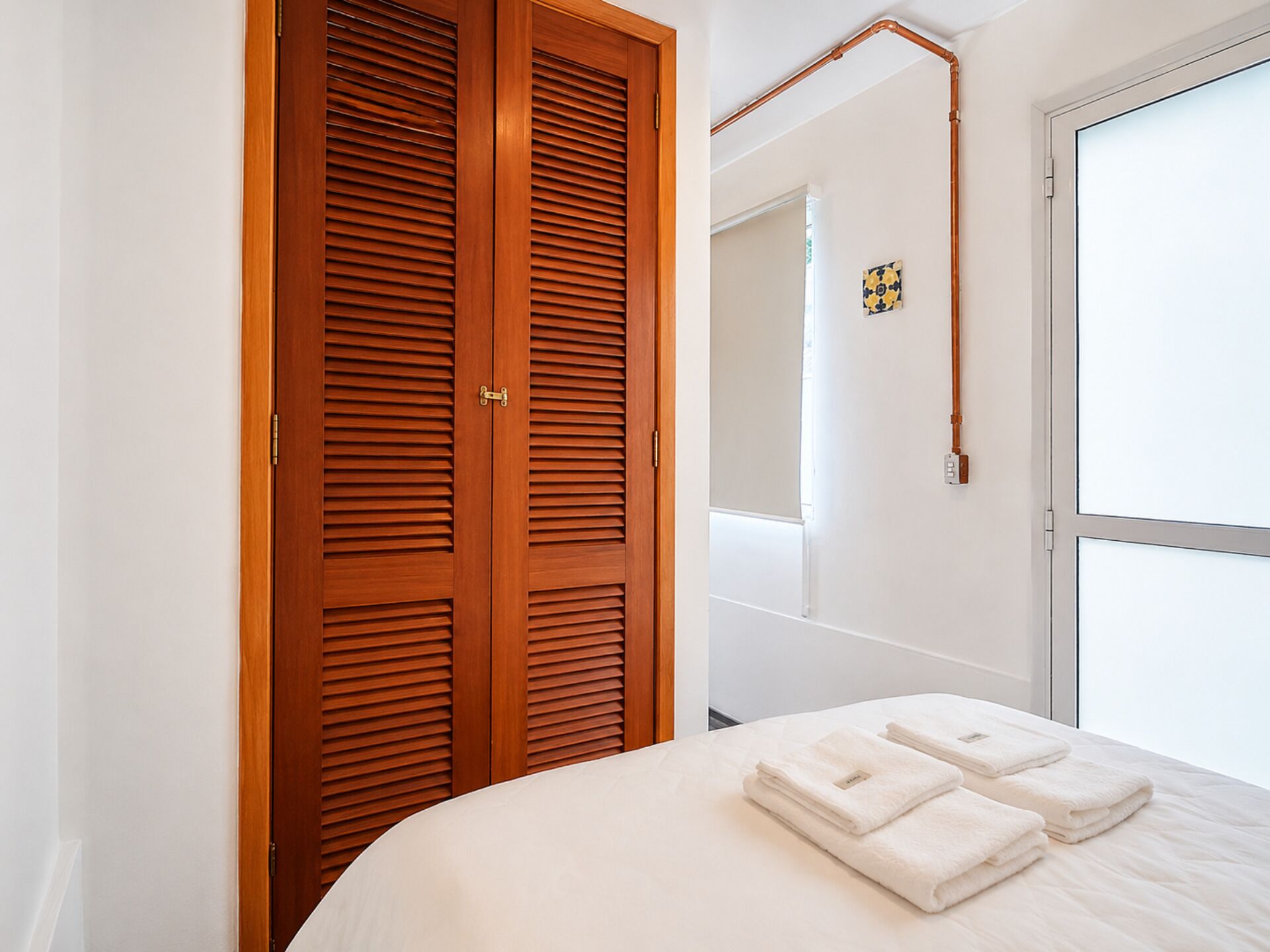 A bedroom with a wooden wardrobe, a bed with white bedding, and a bathroom visible through an open door.