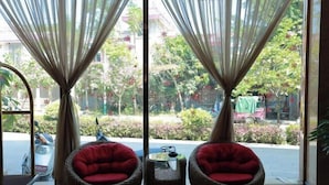 A room with two wicker chairs with red cushions, a small table, and a view of a garden and parked vehicles outside.