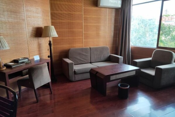 A modern room with a wooden panel wall, a sofa, a coffee table, a desk with a lamp, and a window with a view of greenery.
