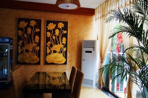 A hotel room with a dining table, two wall-mounted flower paintings, a refrigerator, and a large potted plant. - Mengla Zai Ya Ge Ha Hotel (Mengla)
