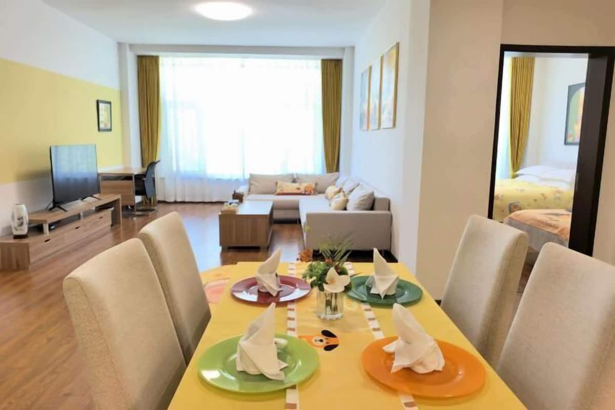 Uma sala de jantar com mesa posta para quatro pessoas, um sofá, uma televisão e uma cama ao fundo.