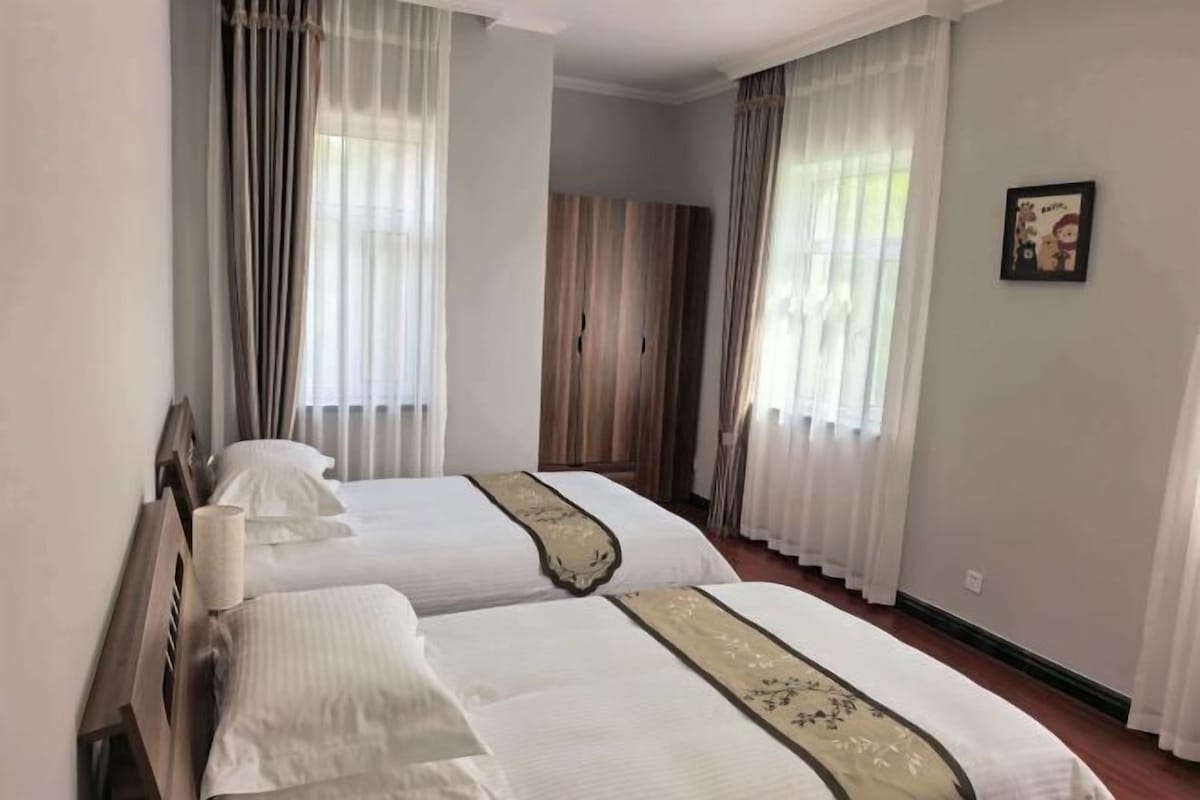 A hotel room with two beds, large windows with curtains, a wardrobe, and a framed picture on the wall.