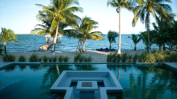 Una piscina con diseño geométrico, rodeada de palmeras y una playa con una estructura de madera.