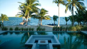 Una piscina amb disseny geomàtric, envoltada de palmeres i una platja amb una estructura de fusta.