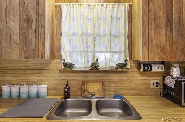 A kitchen with two stainless steel sinks, wooden cabinets, a window with a curtain, and a shelf with kitchen items. - Rustic Lakehouse kayaks fishing bonfires (Inverness)