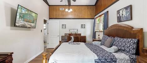 A bedroom with a wooden ceiling, a bed with floral bedding, a ceiling fan, and a TV on the wall.