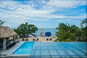 A pool area with a view of the sea, a thatched-roof structure, and lounge chairs. - Conmigos 3 Bedroom Condo - Unit 2 (Placencia)