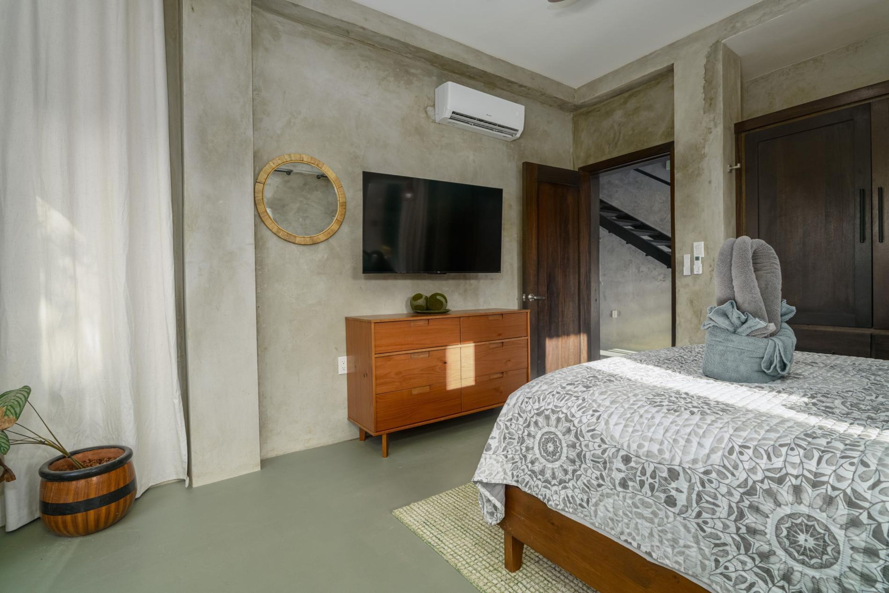Une chambre à coucher comprenant un lit, une commode en bois, un téléviseur, un appareil de climatisation, un miroir, une plante en pot et un escalier.