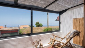 Quarto com uma grande janela, uma cadeira de balanço de madeira, uma mesinha com objetos decorativos e vista para a paisagem.