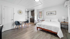 Un dormitorio moderno con una cama grande, un escritorio y una silla. Se observa una zona de cocina al fondo.
