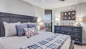 Une chambre à coucher comprenant un lit, une commode, une table de chevet, un ventilateur de plafond et une fenêtre avec des stores.