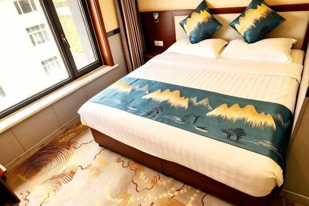 A bed with a mountain landscape bedspread, a window with curtains, and a patterned rug.