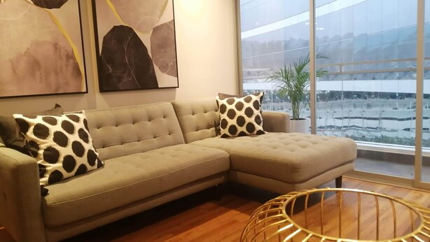 A modern living room with a sectional sofa, abstract wall art, a large window with a view of a cityscape, and a round, wireframe coffee table. - Cozy apartment in Lima, in a club house. (Santiago de Surco)