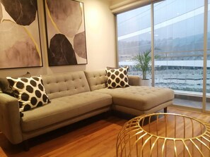 A modern living room with a sectional sofa, abstract wall art, a large window with a view of a cityscape, and a round, wireframe coffee table.