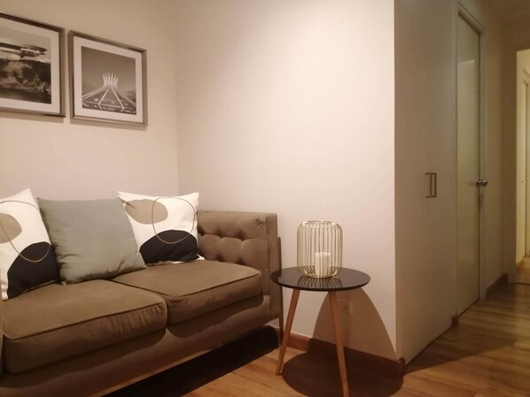 A living room with a brown sofa, two framed pictures on the wall, and a small table with a glass vase.