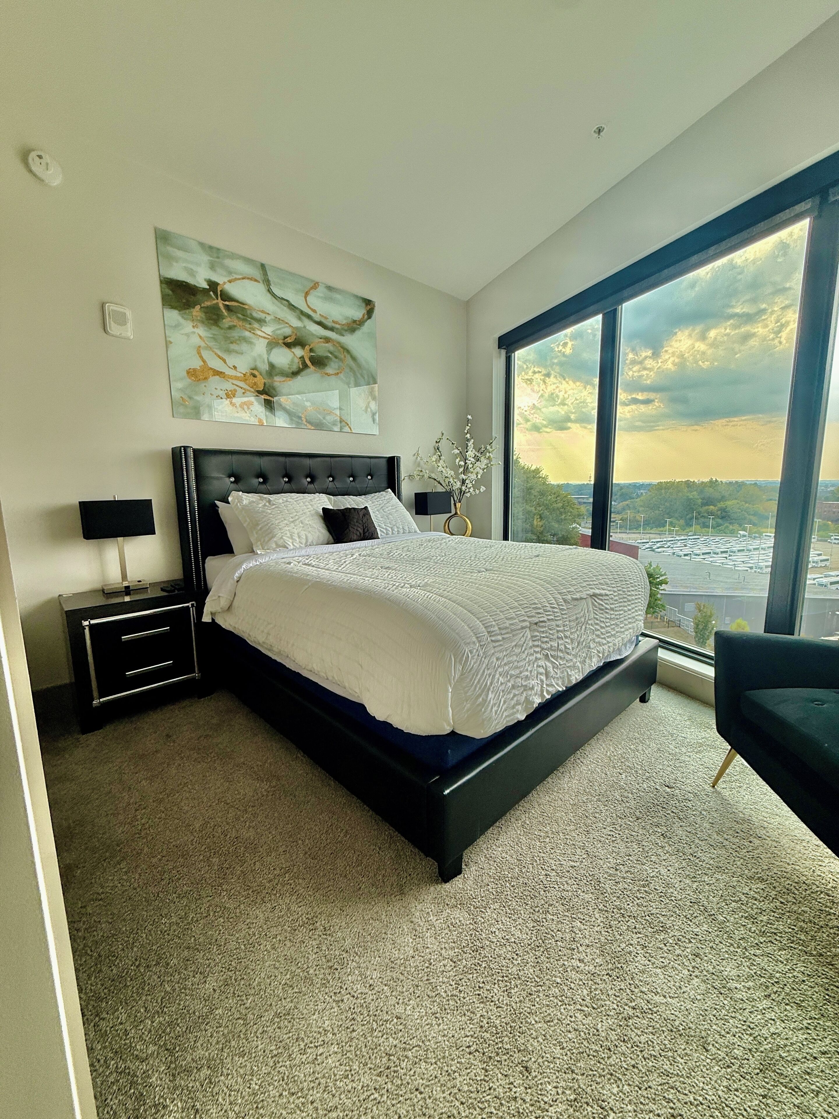 A modern bedroom with a large bed, a nightstand, a lamp, a vase with flowers, and a large window offering a city view.
