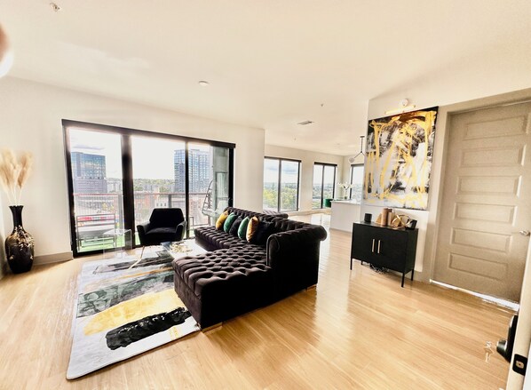A modern living room with a black sofa, a glass coffee table, and a city view through large windows. - Ceiling to floor windows featuring a private balcony with stunning city views!
(Atlanta)