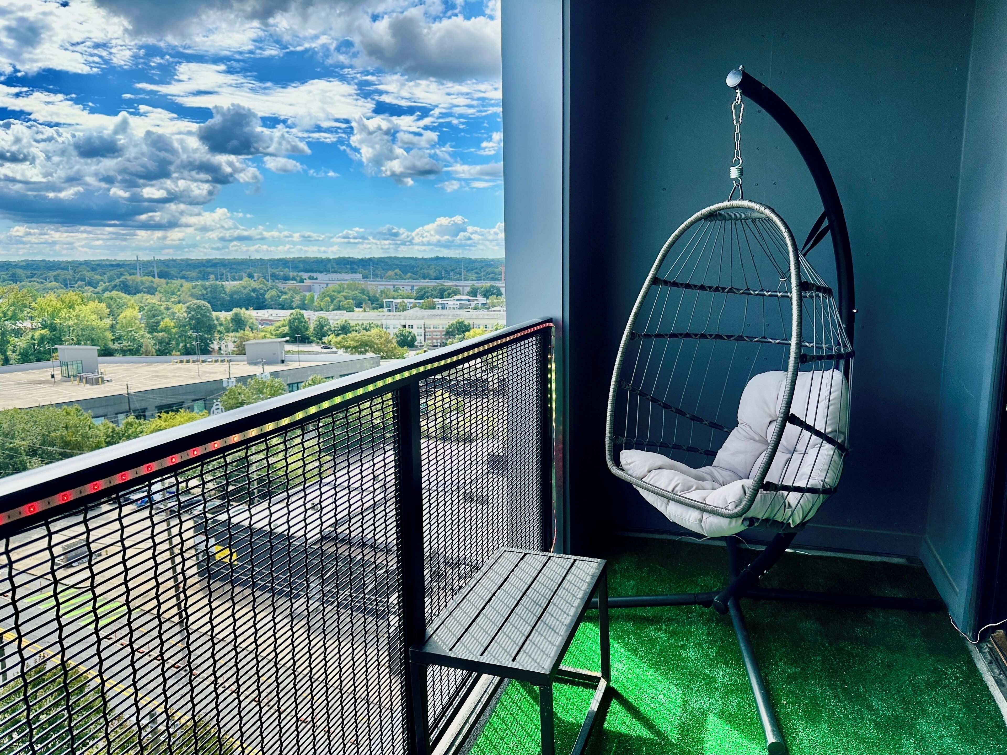 A balcony with a hanging chair, a small table, and a view of a cityscape.
