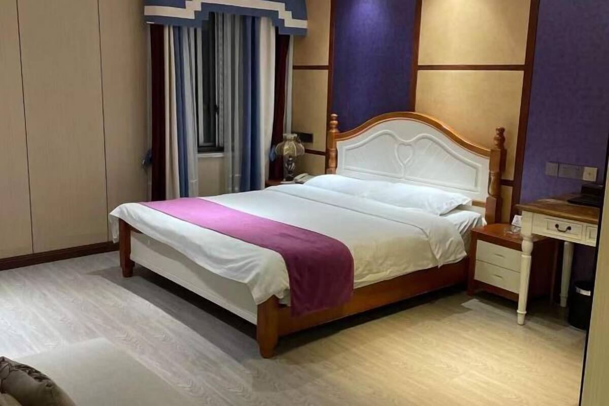 Una cama bien tendida con un cabecero de madera, una mesita de noche con una lámpara y una ventana con cortinas.