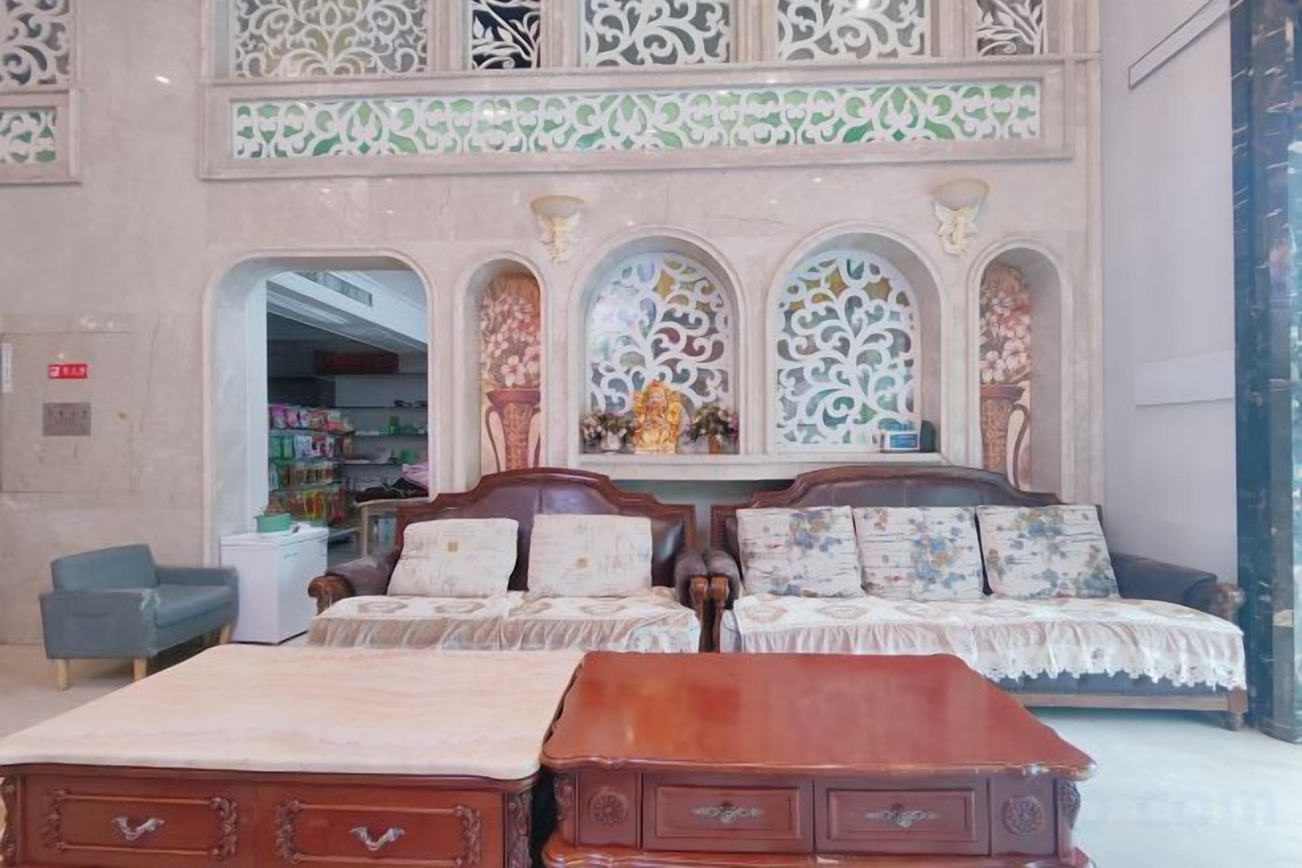 A hotel room with a large bed, a wooden dresser, and a sofa set. There are decorative arches and a chandelier.