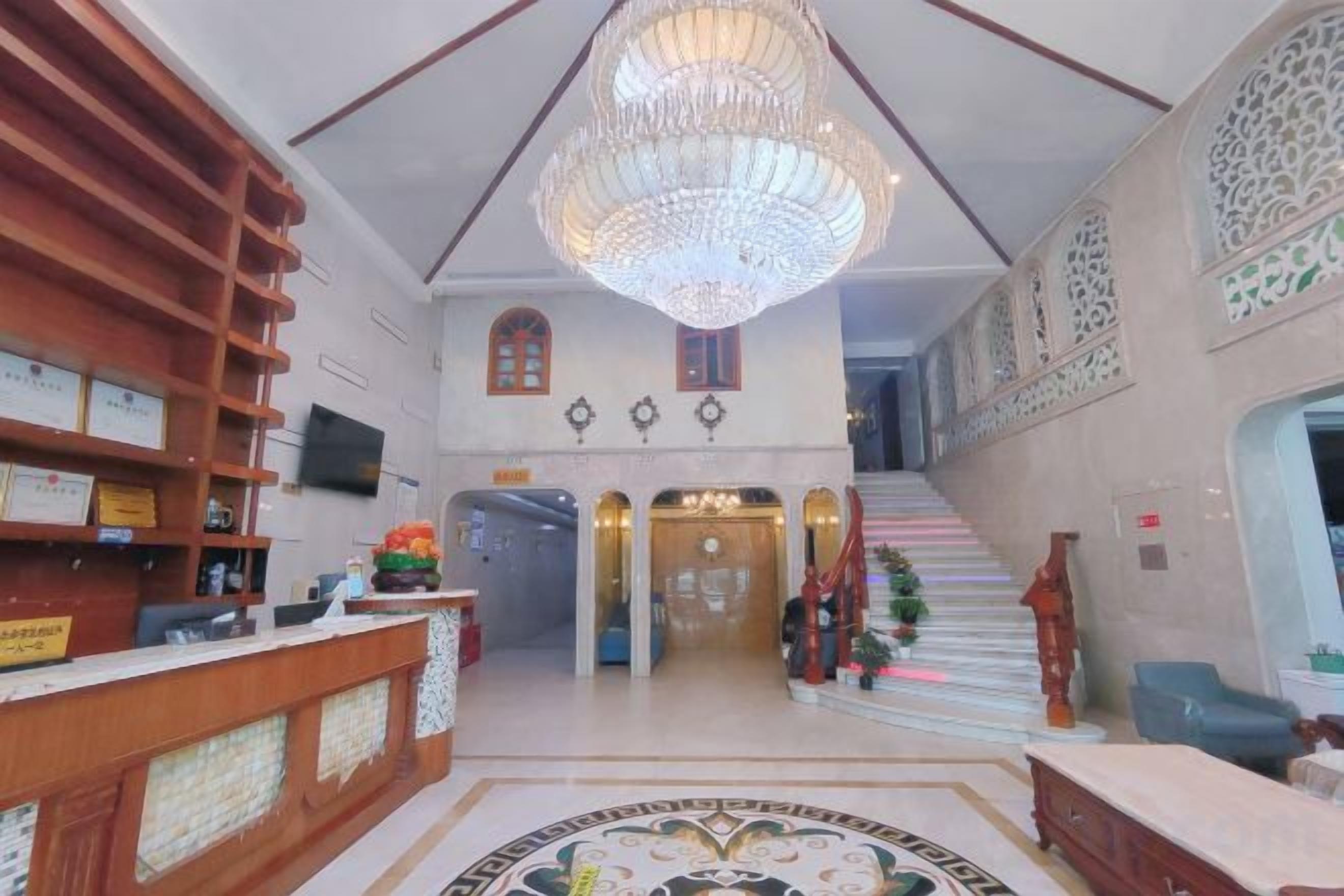 A hotel lobby with a reception desk, a chandelier, a staircase, and a wall-mounted television.