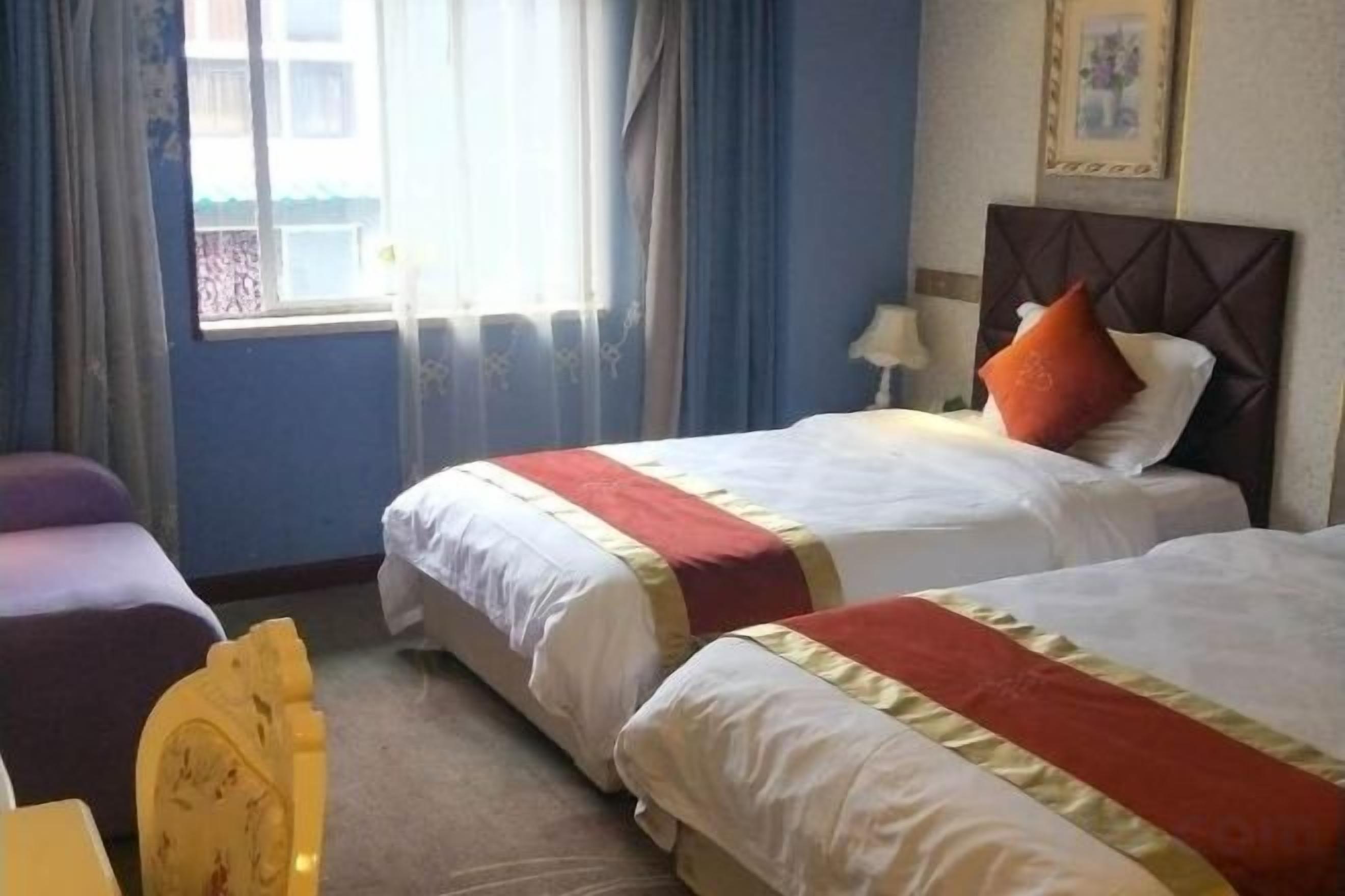 A hotel room with two beds, a window with curtains, a bedside table with a lamp, and a framed picture on the wall.