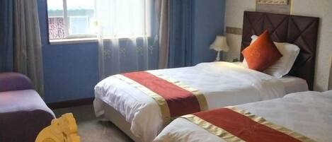 A hotel room with two beds, a window with curtains, a bedside table with a lamp, and a framed picture on the wall.