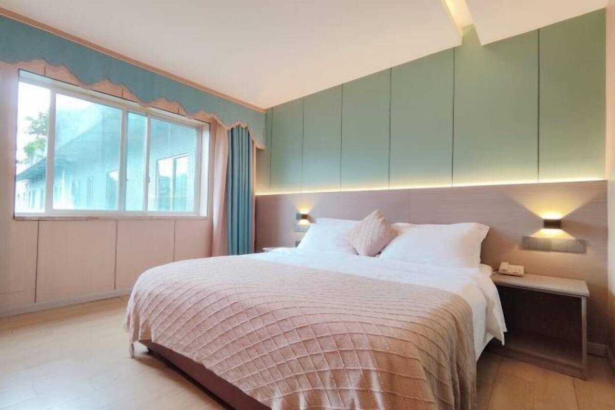 Un dormitorio con una cama grande, una mesita de noche, una ventana con cortinas y una lámpara fijada a la pared.