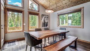 Un comedor con mesa y bancos de madera, ventanas amplias con vistas al bosque y techo con vigas de madera a la vista.