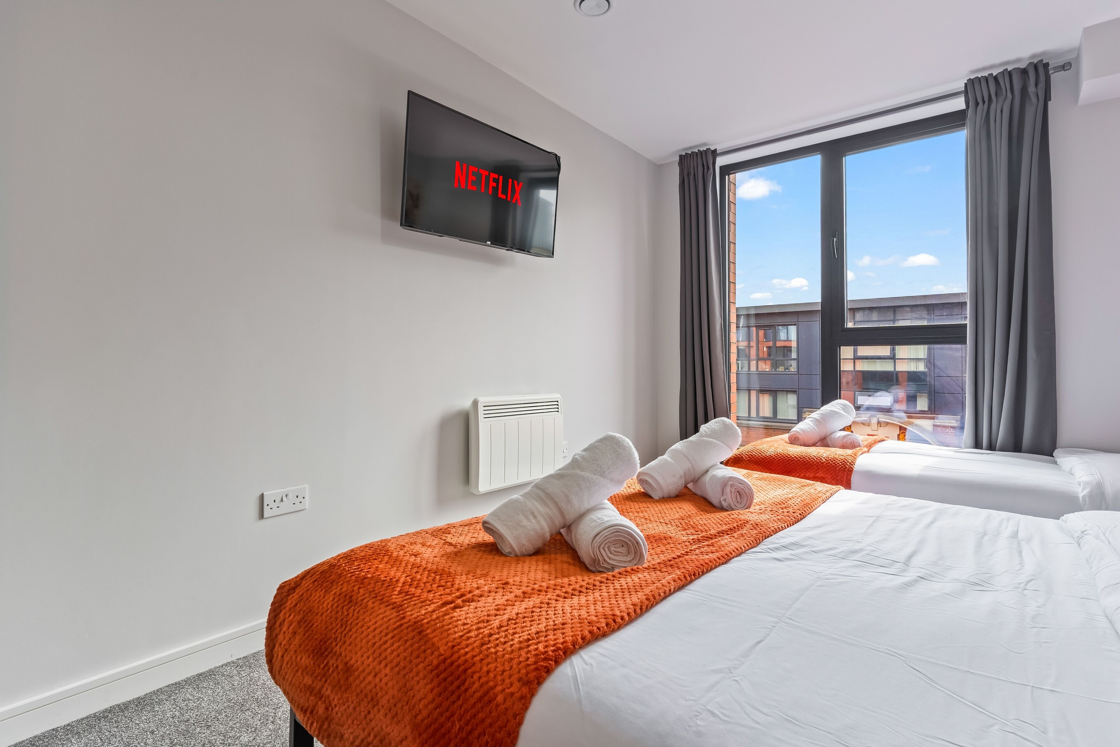Habitación de hotel con dos camas, un televisor mostrando 'NETFLIX', una ventana con cortinas y vistas a edificios.