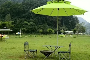 A grassy field with a table and chairs under a green umbrella, surrounded by tents and trees.