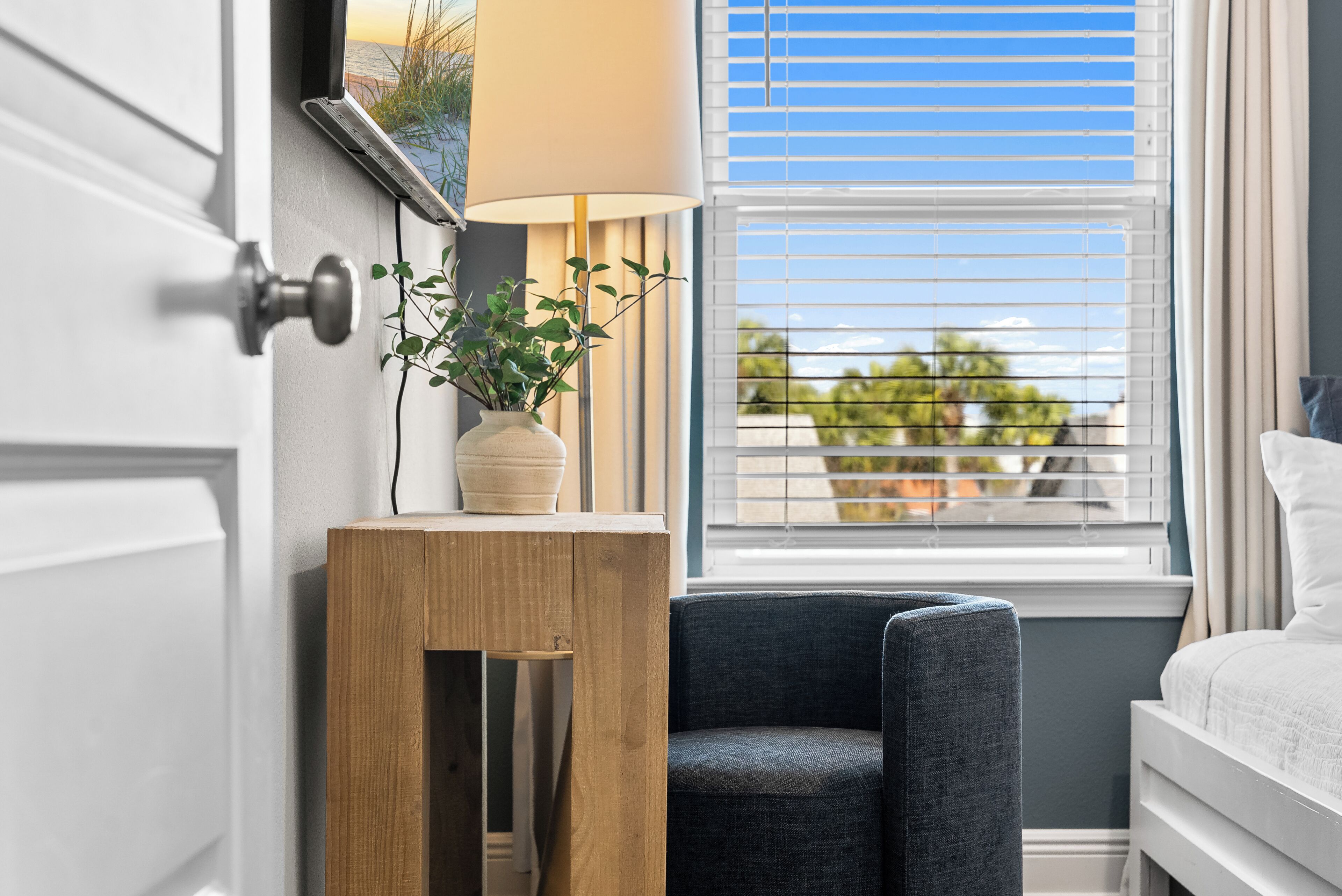 A bedroom with a bed, a window with blinds, a lamp, a small wooden table, and a chair.