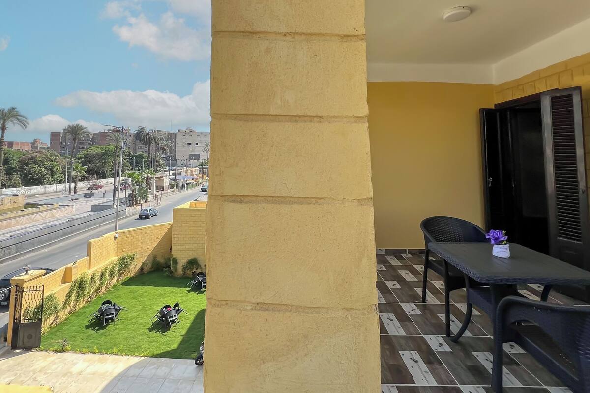 A balcony with a view of a road and palm trees, a table with a chair, and a small plant on the table.