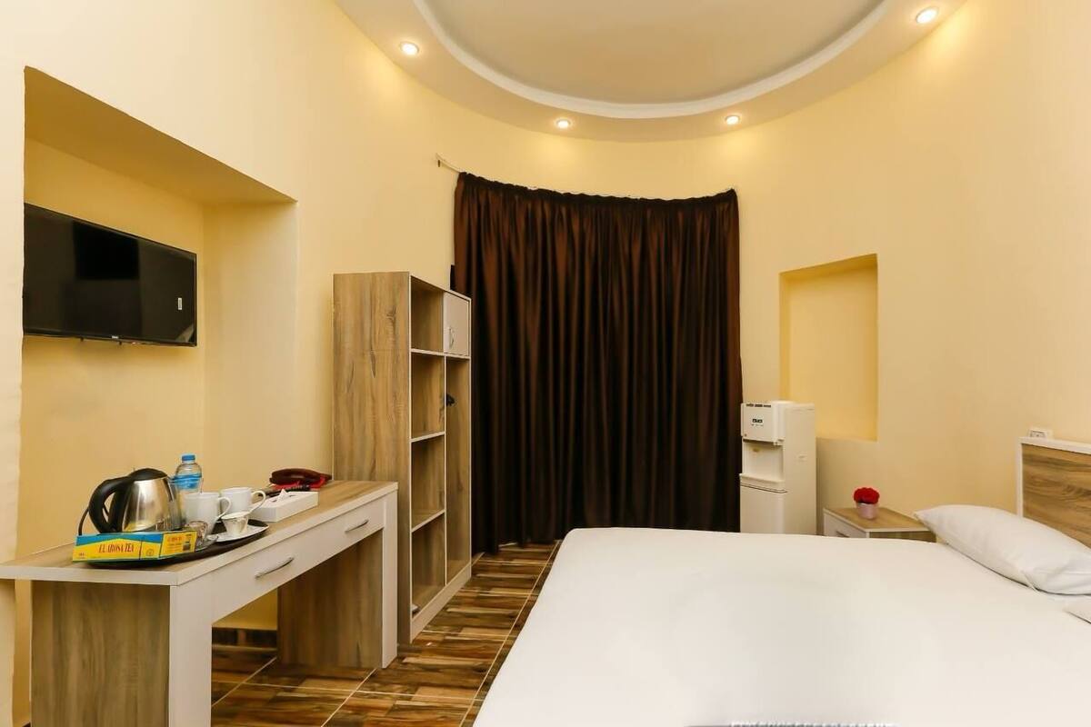 A hotel room with a bed, a desk with a flat-screen TV, a wardrobe, a bedside table with a lamp, and a curtain.