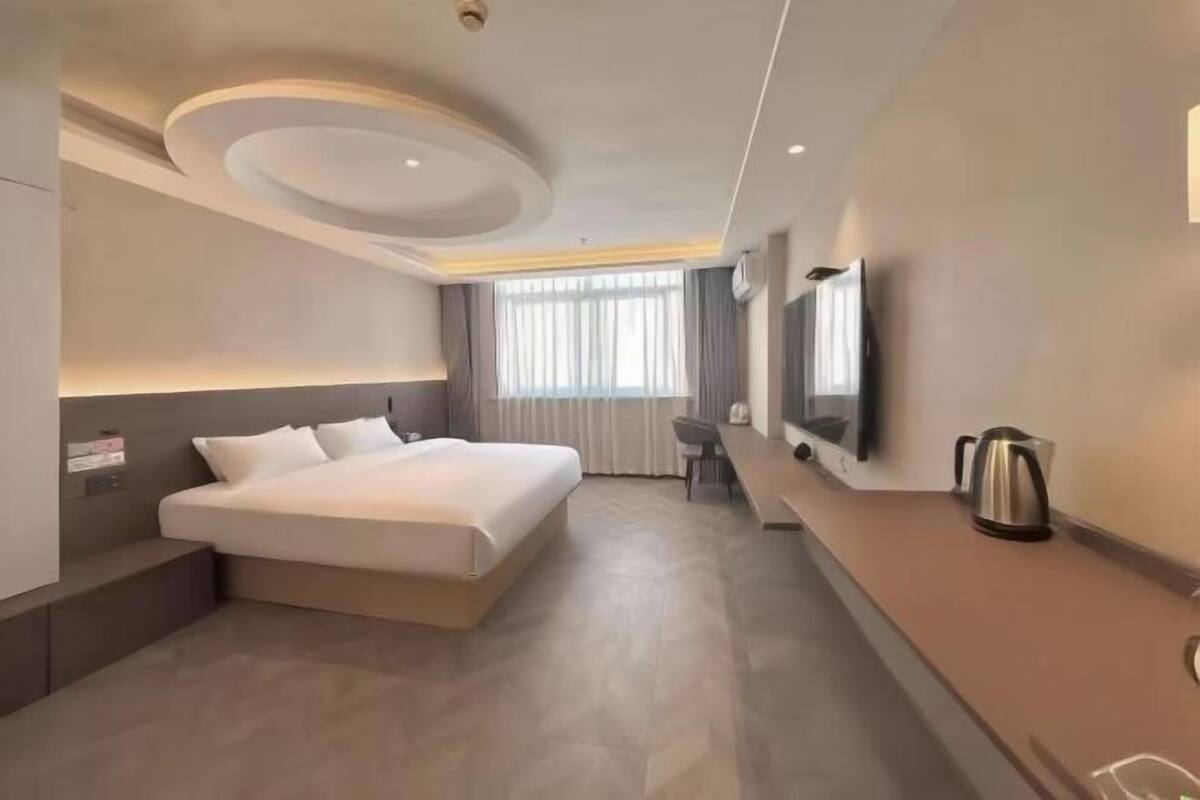 Ein modernes Hotelzimmer mit einem großen Bett, einem Schreibtisch, einem Sessel, einem Fernseher und einem Wasserkocher auf der Ablagefläche.