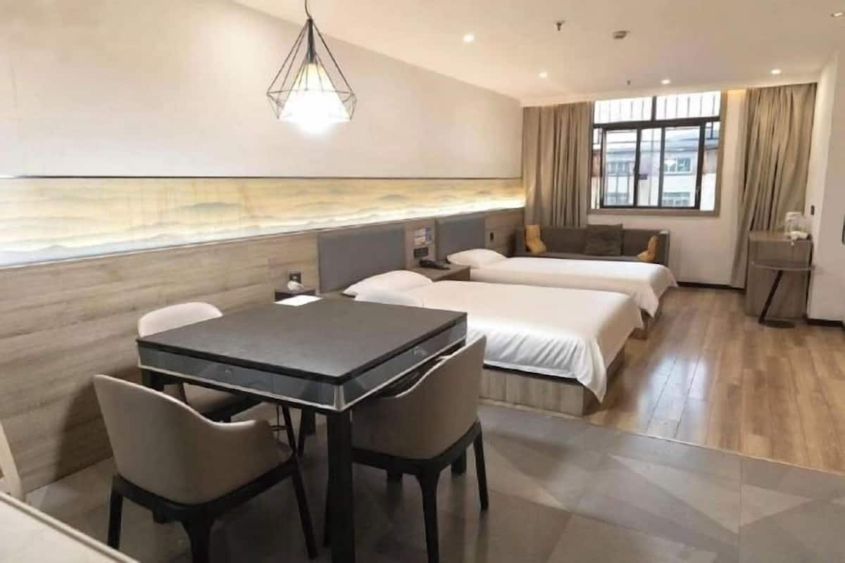 Ein Hotelzimmer mit Esstisch, zwei Betten, einem Nachttisch, einer Lampe und einem Fenster mit Vorhängen.