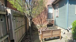 Un jardin à l’arrière d’une maison, avec une clôture en bois, un banc, un petit arbre et une maison à l’extérieur verdoyante.