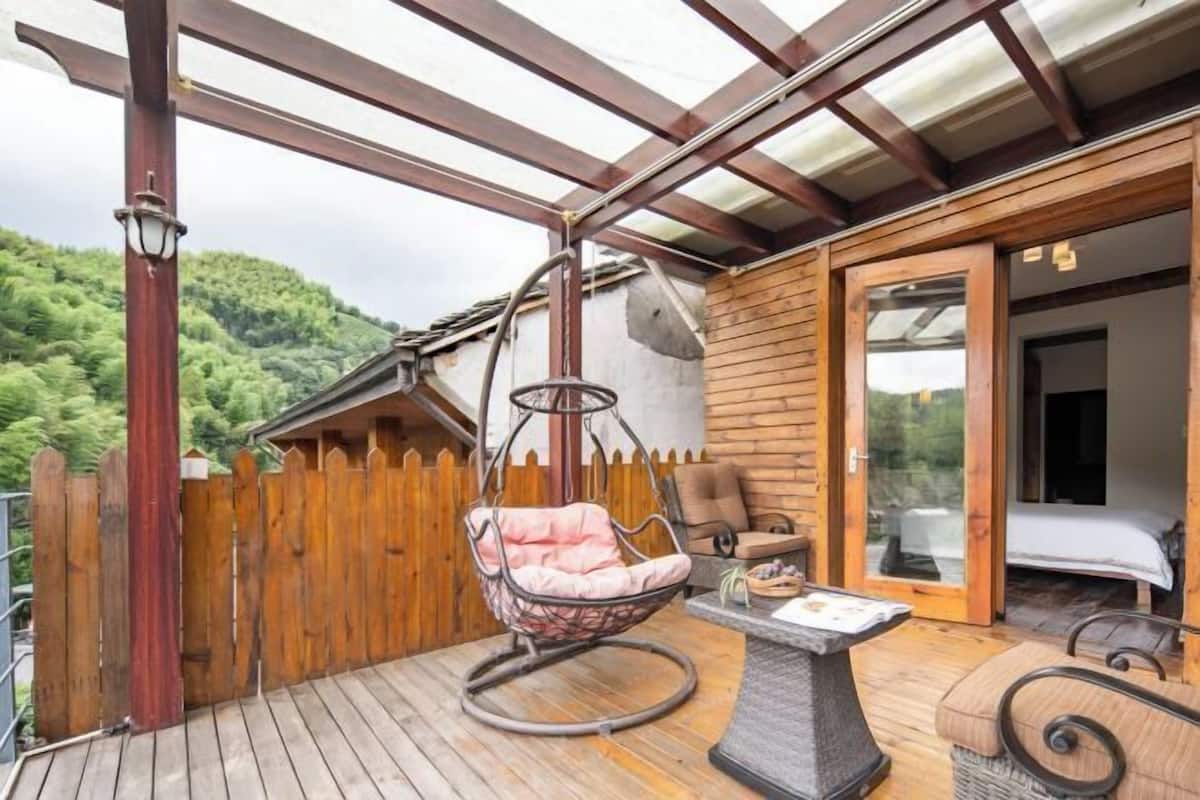 Una terraza de madera con una hamaca colgante, una mesa y una cama en una habitación con vistas a una colina boscosa.