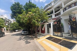 Une rue résidentielle bordée d’arbres, avec un trottoir, une entrée fermée par un portail et une voiture garée.