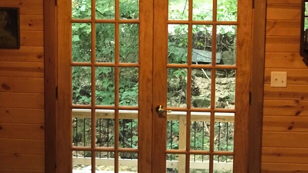 A wooden double door with a grid pattern, leading to an outdoor area with a wooden deck and greenery.