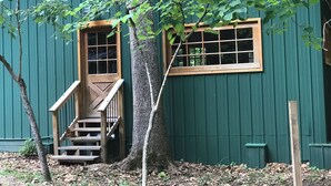 A green building with a wooden staircase leading to a door, a window, and a tree in front.