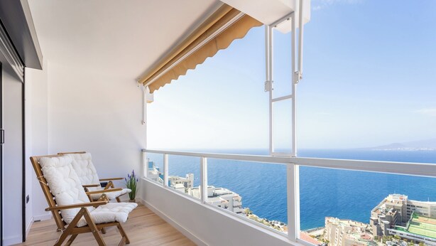 Ein Balkon mit Blick auf das Meer und Gebäude, Holzstühle und ein Blumentopf.