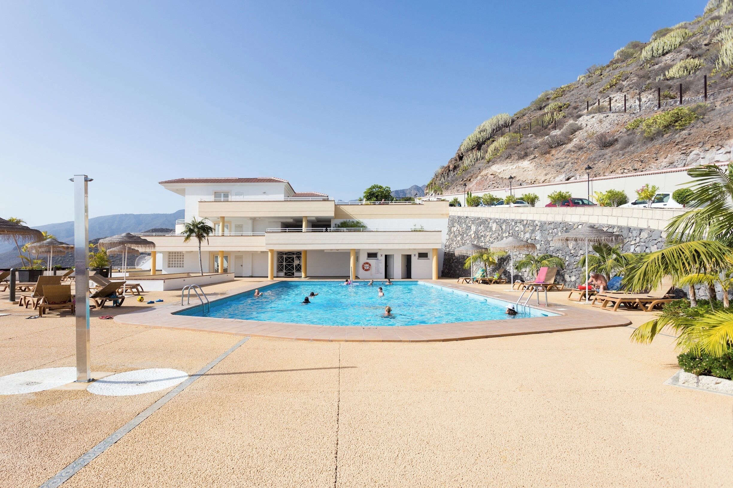 A pool area with a large pool, lounge chairs, and a building with a flat roof.