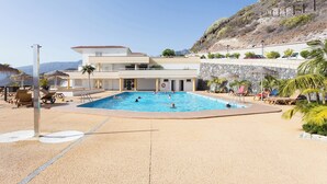A pool area with a large pool, lounge chairs, and a building with a flat roof. - Home2Book Sunset Apt Costa Adeje, Pool & Terrace (Adeje)