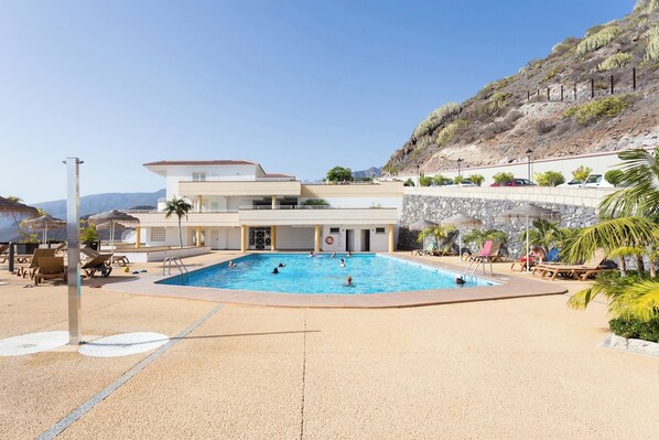 A pool area with a large pool, lounge chairs, and a building with a flat roof.