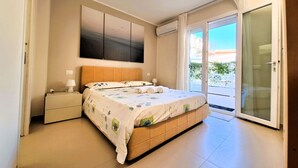 A bedroom with a bed, a nightstand, a large window, and a sliding glass door leading to a balcony.